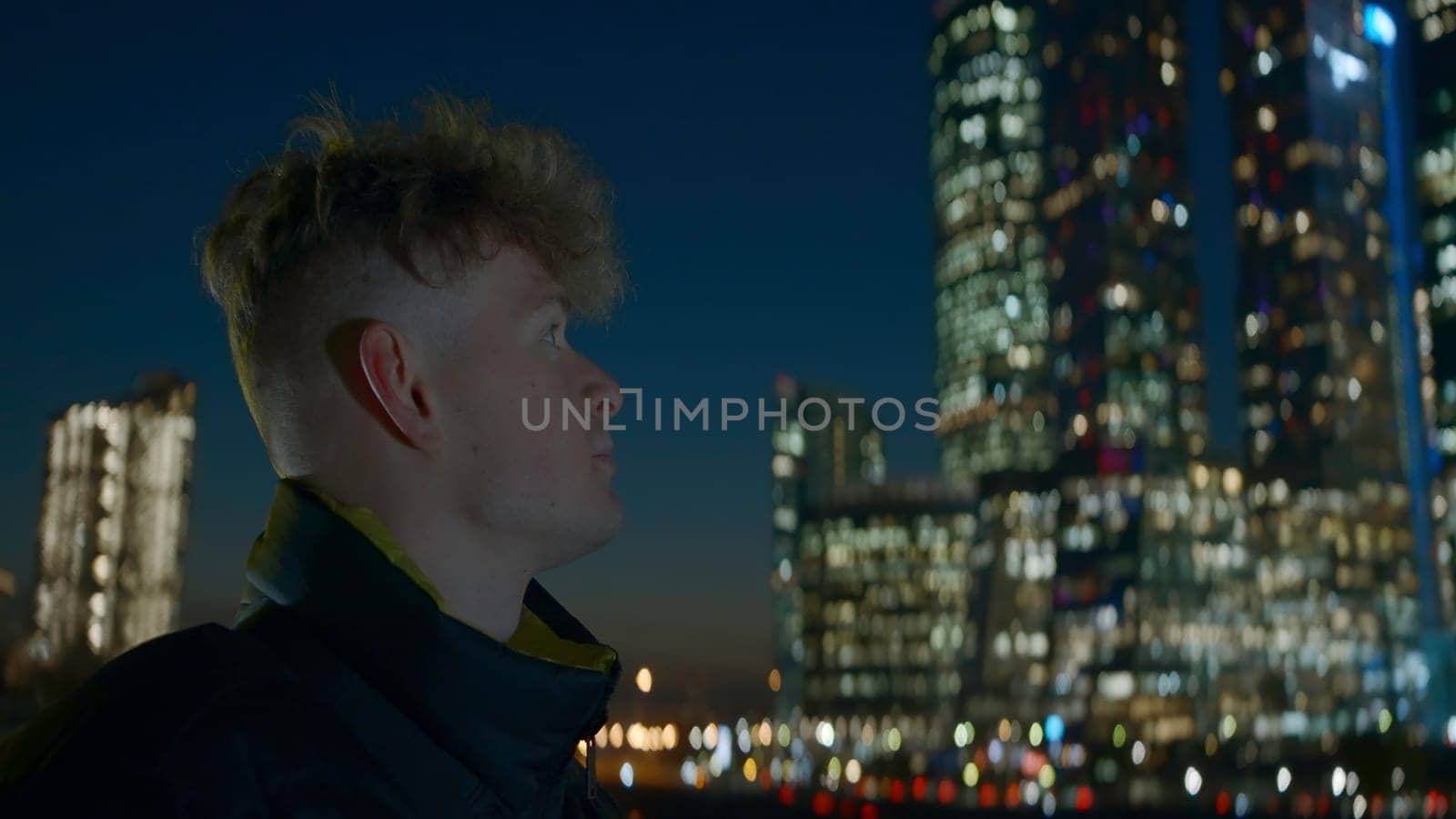 Man looks at modern glowing skyscrapers at night. Action. Young man on background of bright glowing skyscrapers at night. Man looks at skyscrapers glowing at night in city.