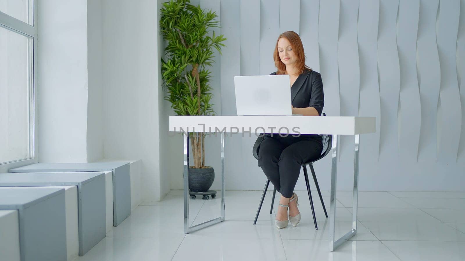 Working woman with red hair. Action.A woman in a black suit in a cozy room who is sitting at a computer desk in a laptop working. High quality 4k footage