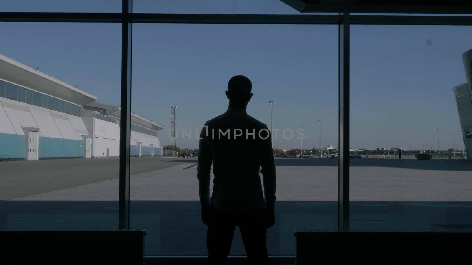 Back view of a confident successful male silhouette in front of a large window. HDR. Thoughtful businessman watching the summer blue sky and a parking space