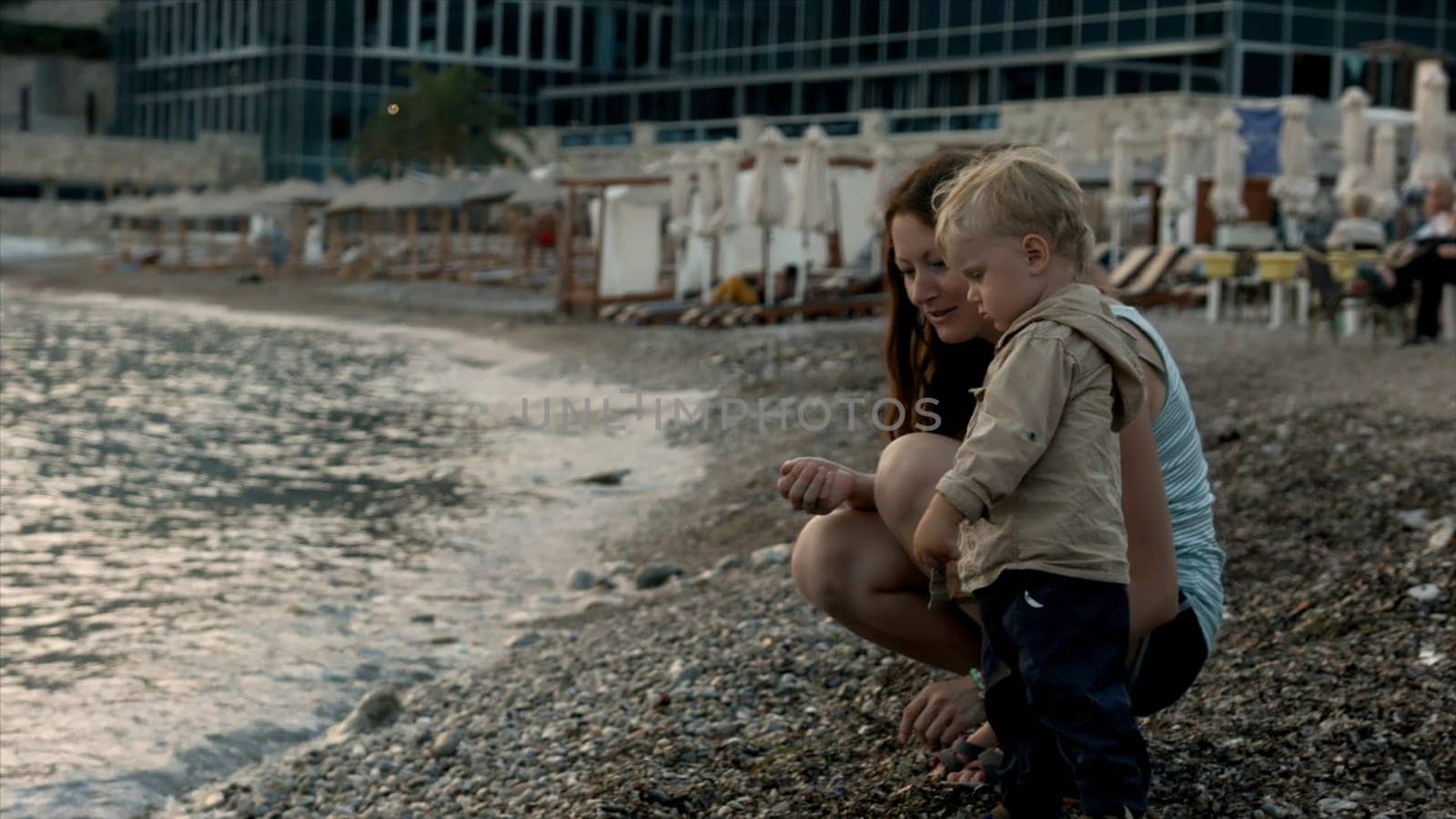 A young mother and her son throw stones in rippling water. Creative. Little boy with mom throw pebbles into the sea. by Mediawhalestock