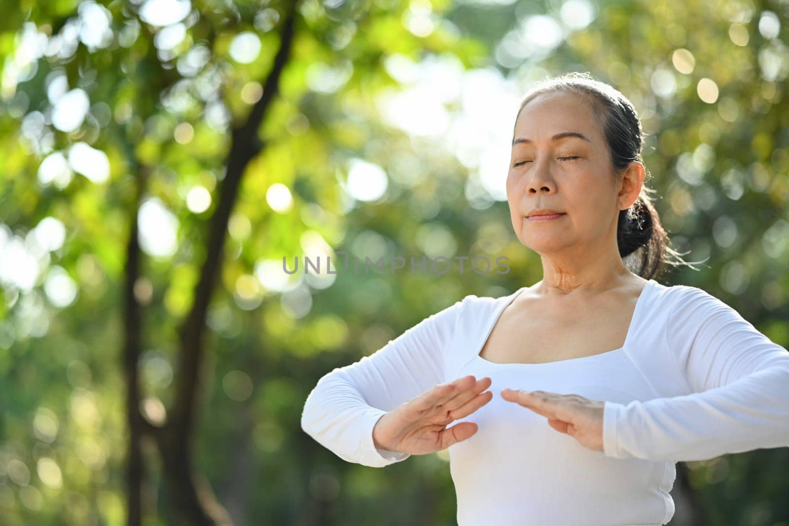 Calm senior woman with eyes closed meditating or practicing traditional Tai Chi Chuan in nature. by prathanchorruangsak