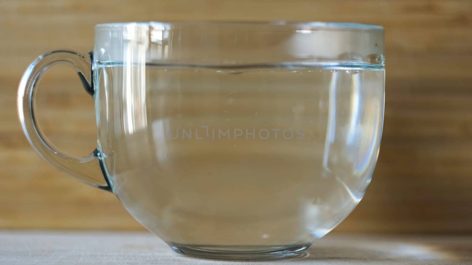 Pouring hot water into a big transparent cup on blurred wooden background. Side view of an empty cup and the process of poring hot water for the morning hot beverage.