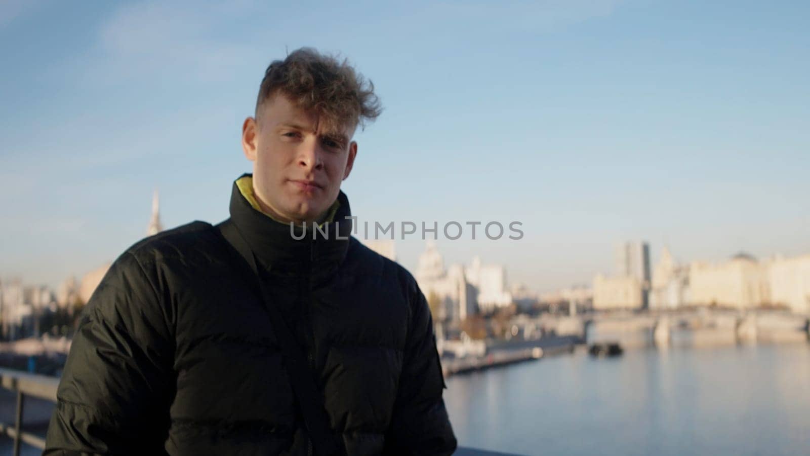 Man with short curly hair walking around the city. Action. Concept of travelling, man in wark jacket walking with the river on the background