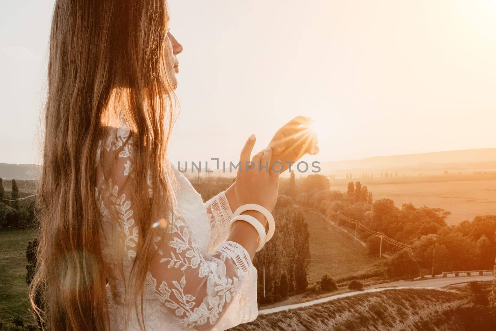 Happy woman in white boho dress on sunset in mountains. Romantic woman with long hair standing with her back on the sunset in nature in summer with open hands. Silhouette. Nature. Sunset. by panophotograph