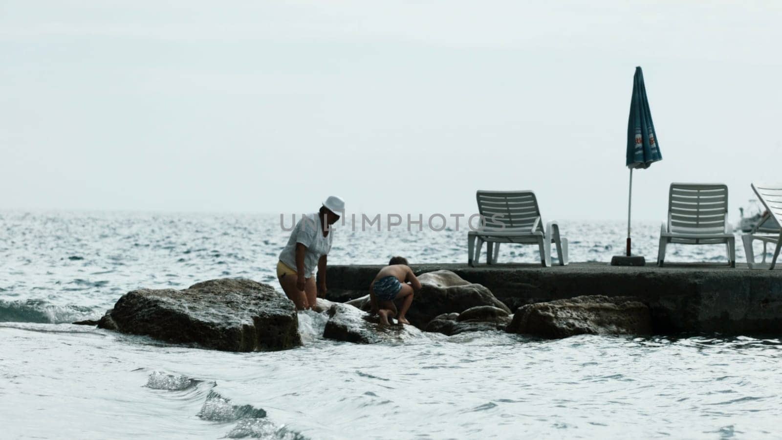 Grandmother and a boy child playing by the sea and stones. Creative. Spending time by the pier on summer vacation. by Mediawhalestock