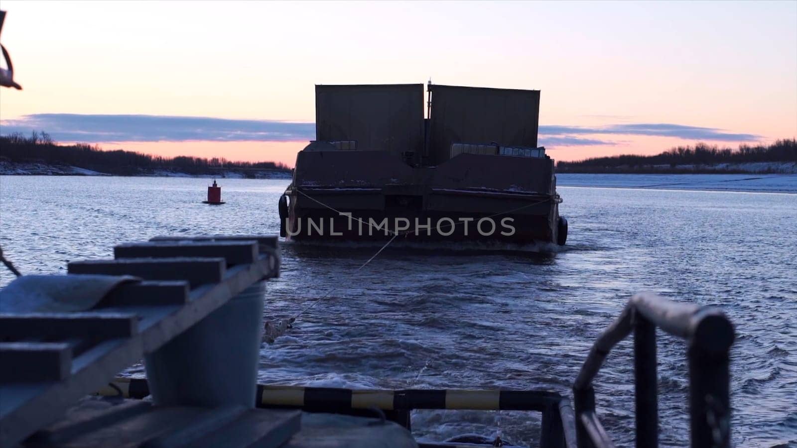 Barge in tow of fishing boat. Clip. Boat carries barge with cargo in cloudy twilight. Boat and barge float on river on winter morning.
