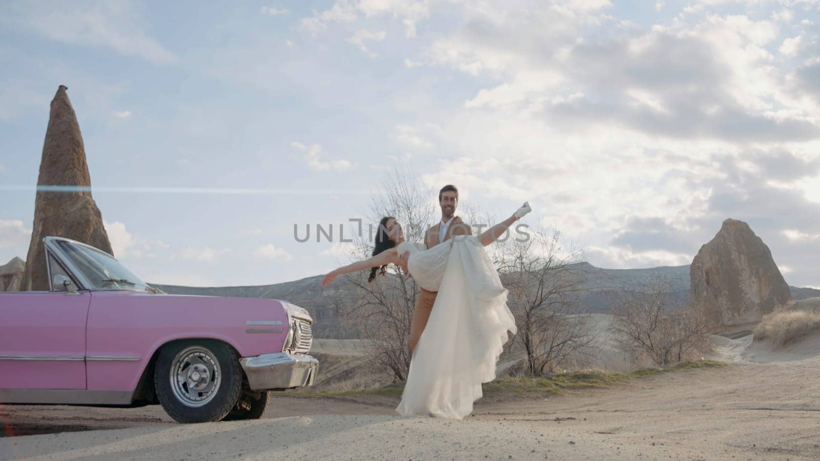 Happy wedding couple in mountains posing in front of a camera. Action. Groom in suit holding beautiful bride in his arms near the cabriolet pink car