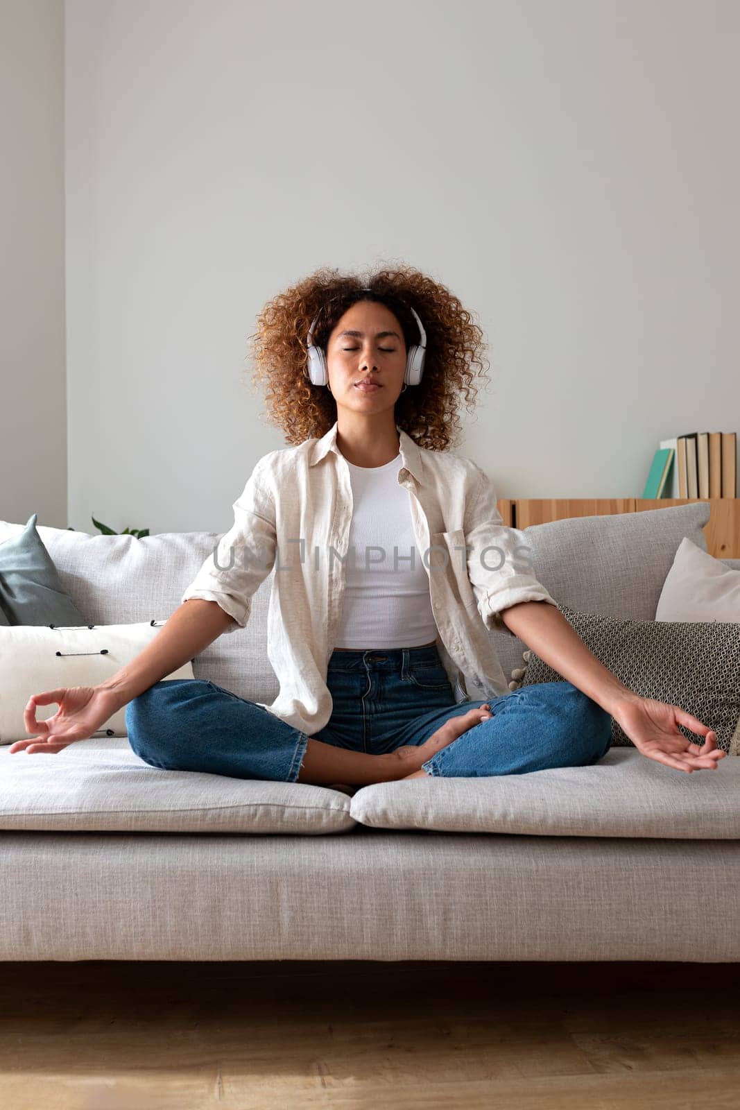 Front view vertical portrait of multiracial hispanic young woman meditating on the couch listening to guided meditation using wireless headphones. Technology and mental health concept.