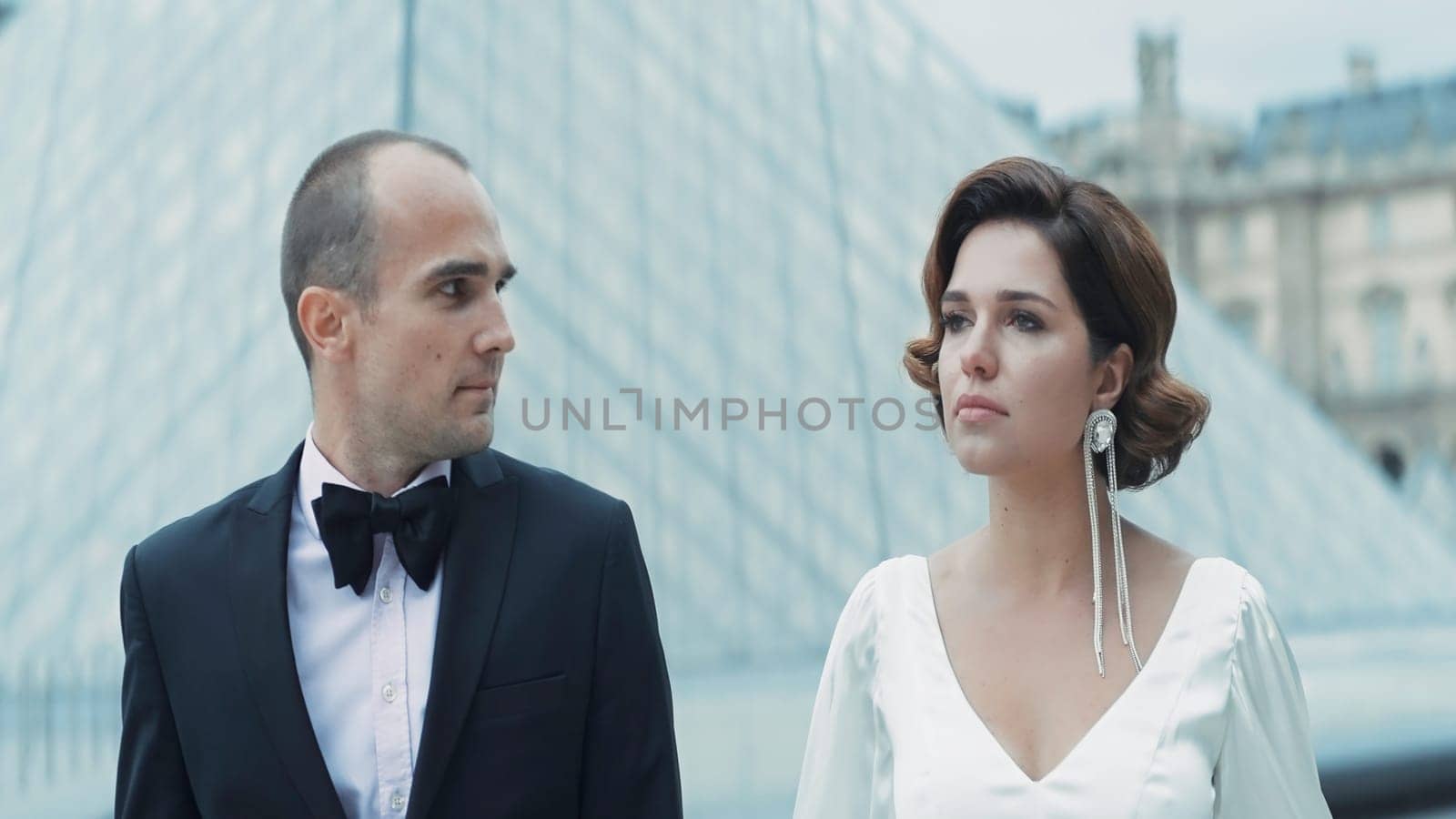 Beautiful elegant couple of lovers walking in ancient city. Action. Luxurious couple walks in love in Paris. Couple in elegant outfits walks on background of glass pyramid by Mediawhalestock