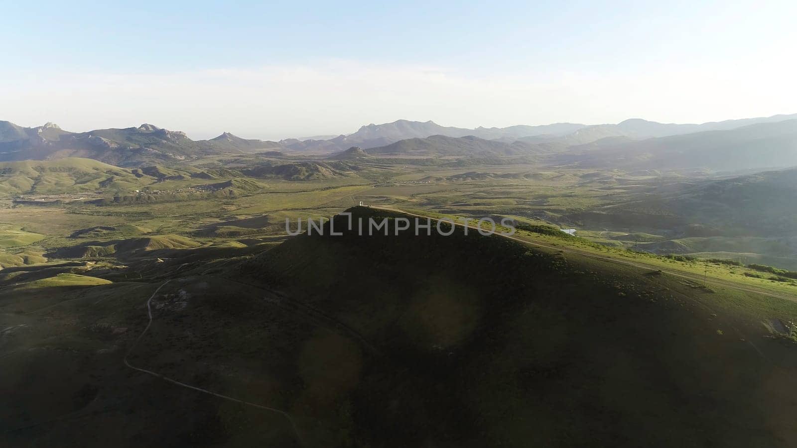 Aerial view of country road on the top of high green hill with meadows and mountain chain on the background against blue sky. Beautiful view from above