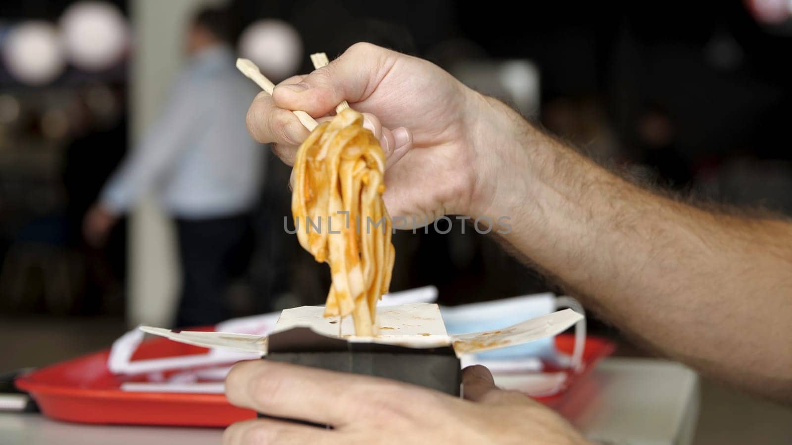 Close up of male hands with wooden chinese chopsticks eating delicious asian wok noodles. Side view of eating chinese pasta in a public place.
