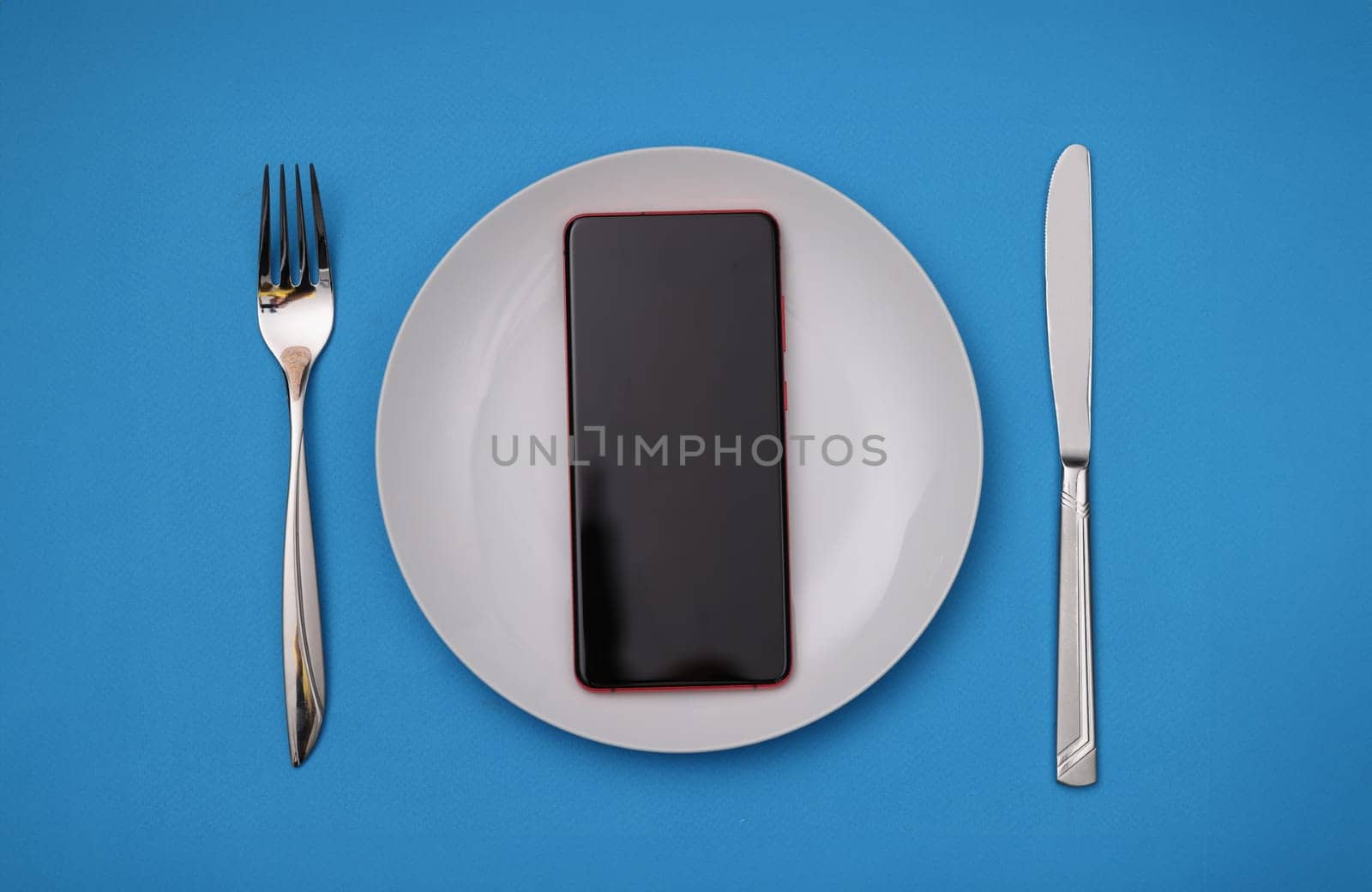 Mobile phone lying in white plate near fork and knife. Phone addiction concept
