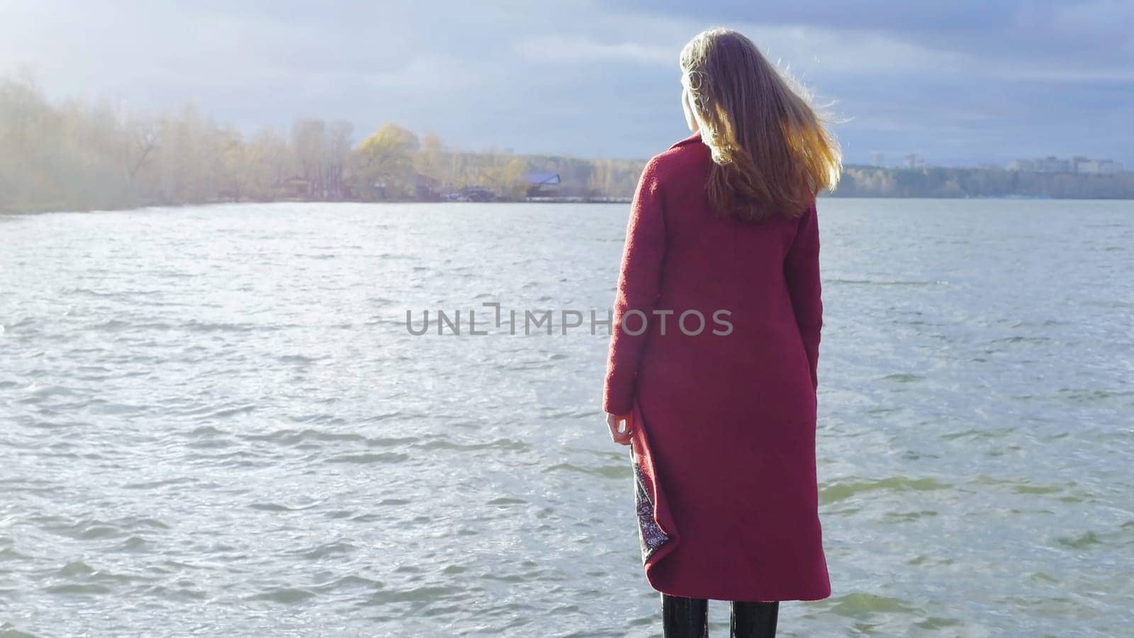 Young girl with her hair stands near the water in the cold season. Back view. Young woman standing near a lake.