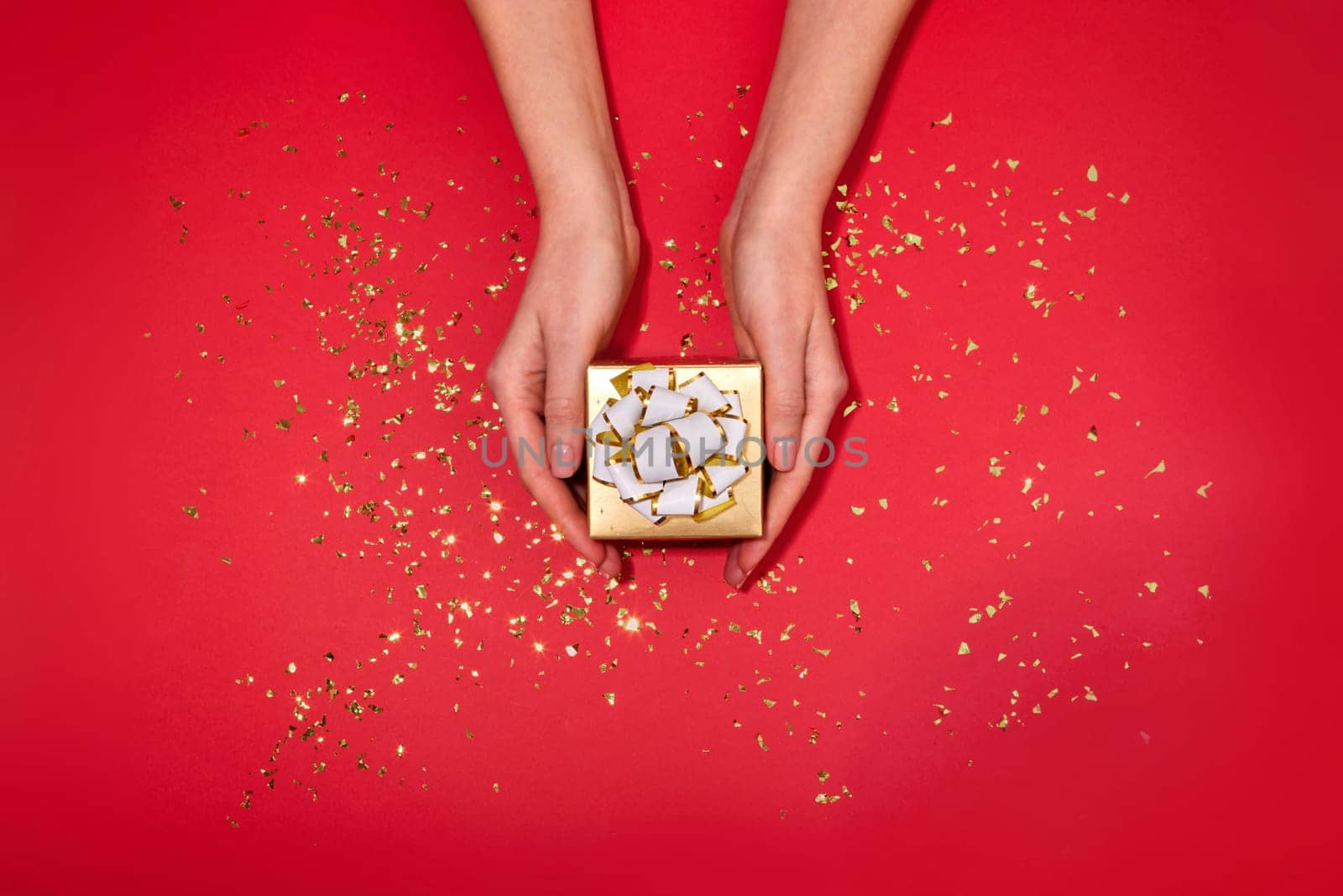 Female hands holding small gold box with a white bow gift hidden inside on red maroon background with scattering of gold sparkles and glitter top view Christmas New Year Valentine's day birthday
