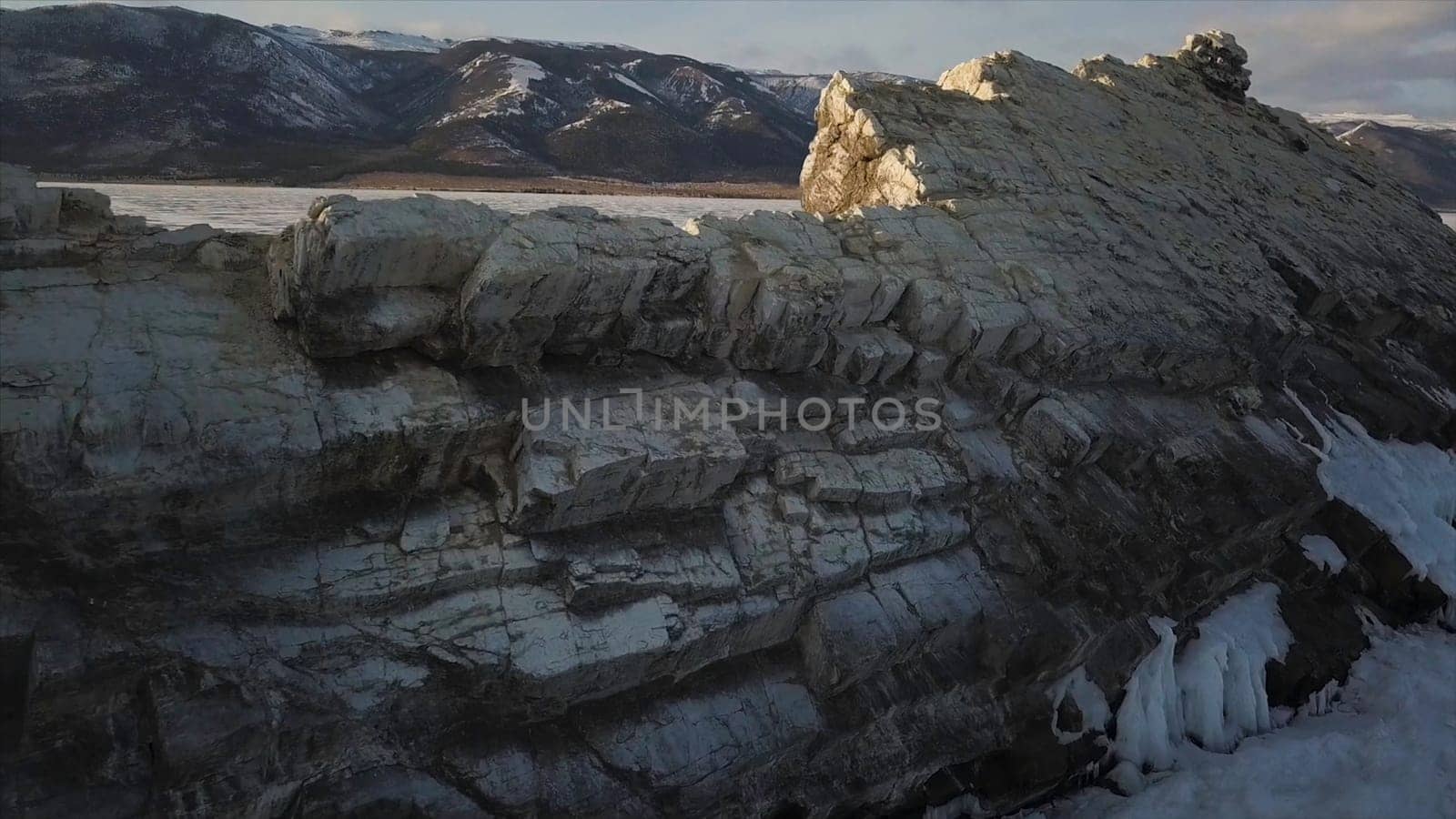 Aerial view of the cliff with steep slopes on the background of forested mountains and frozen pond. Mountains with peaks partially covered with snow by the beautiful frozen Baikal Lake under blue sky.