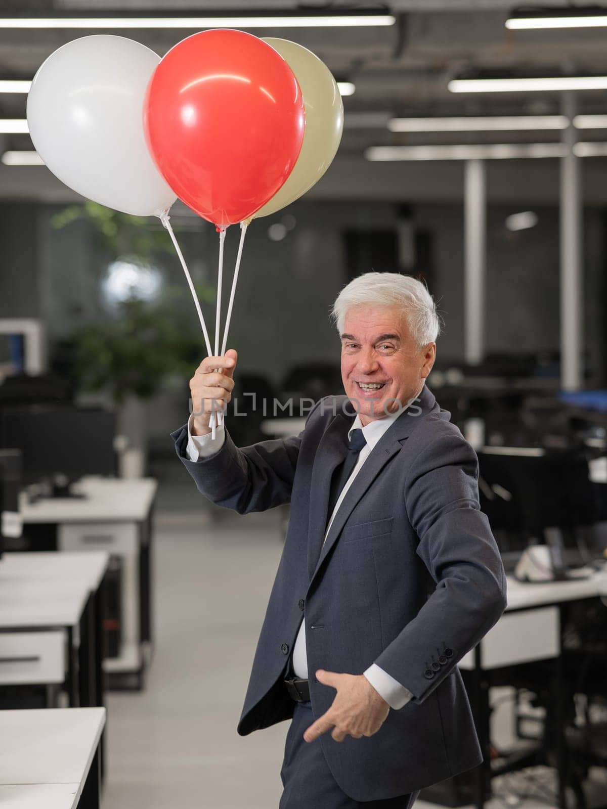 Portrait of a cheerful mature business man holding balloons in the office. Vertical photo