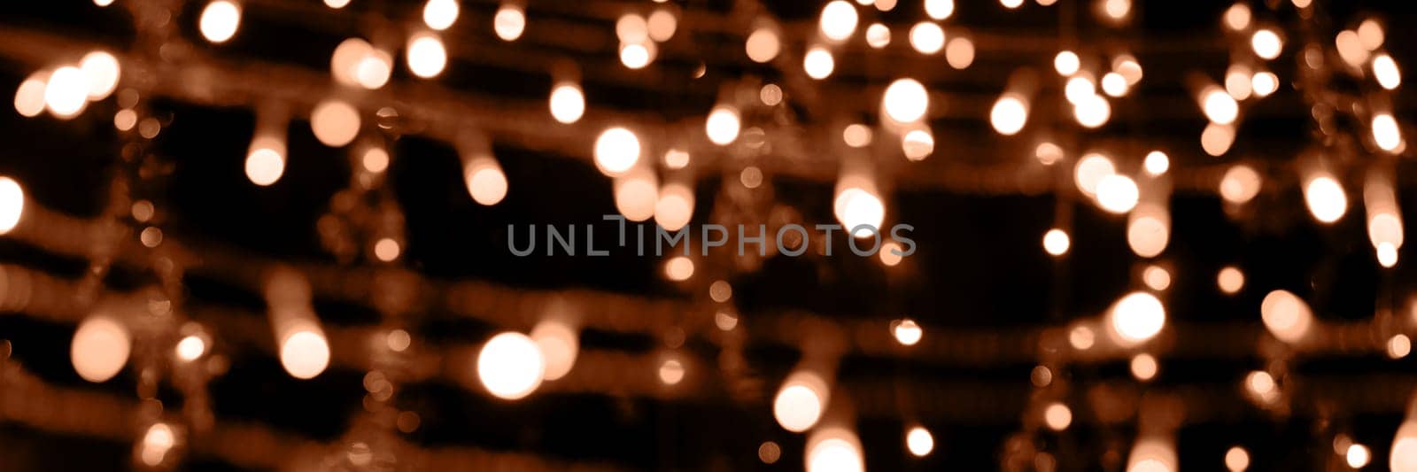 Blurry garland lights on a dark background. Festive Christmas and New Year background. Soft focus. Image toned in color of the year 2024 - Peach Fuzz.