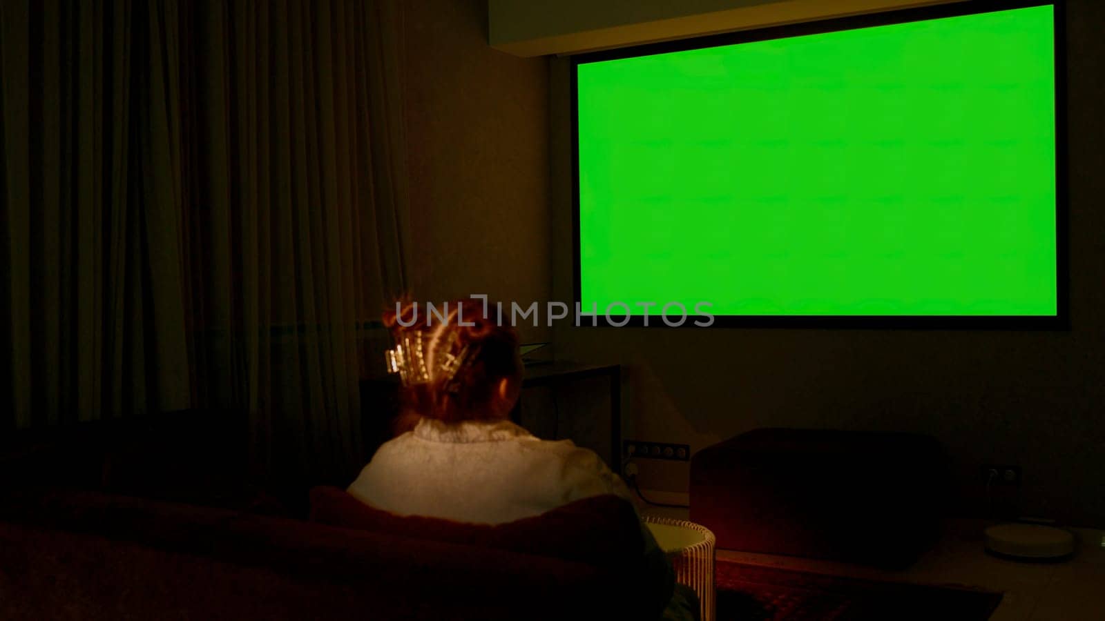 Woman watching television in living room late in the evening. Media. Young girl sitting on sofa against tv with chroma key green screen, back view