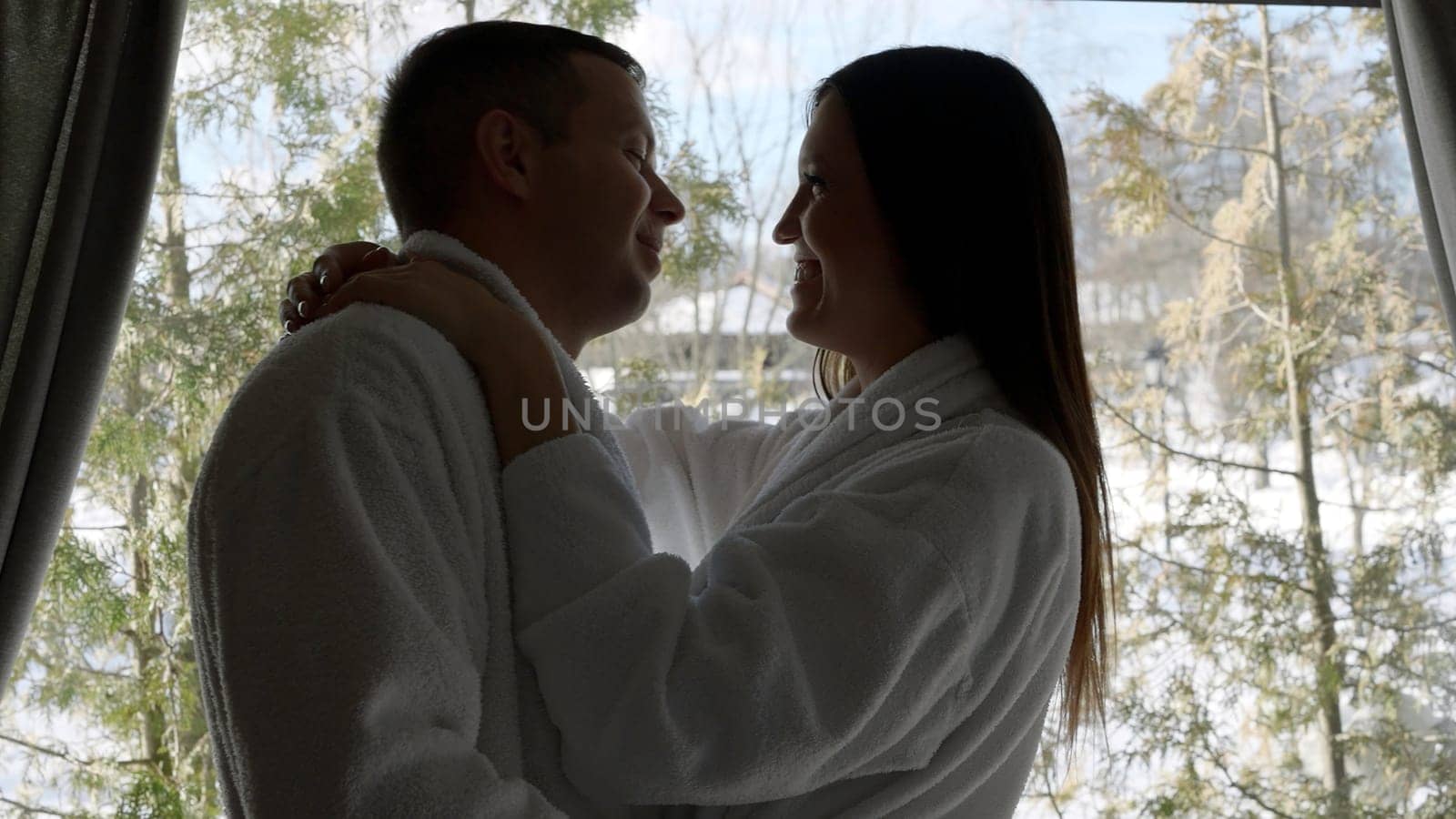 Loving man and woman are hugging in morning in bedroom against window. Action. Husband and wife near window with trees behind