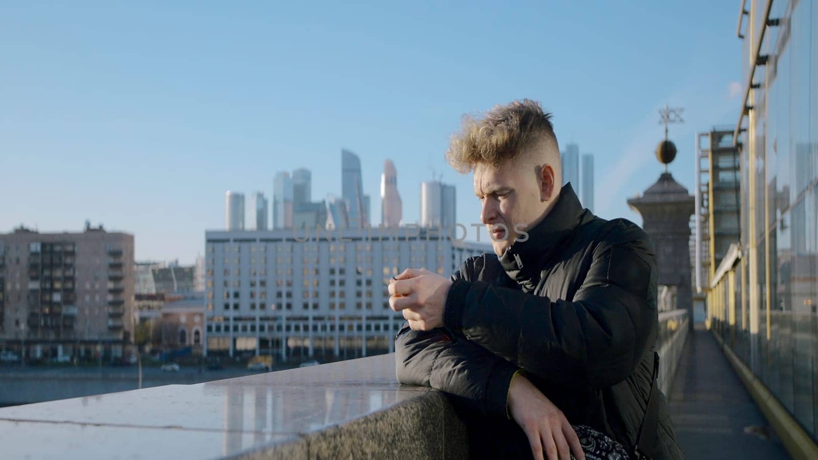 Young man looks at city from bridge. Action. Man on bridge overlooking sunny modern city. Man in jacket stands on bridge on sunny day.