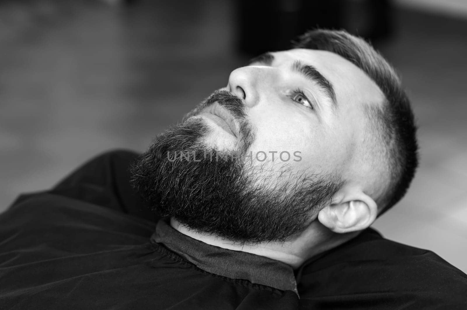 A young man with a beard in a black cape sits on a chair in a barbershop. The client is waiting for a haircut of the beard. by Niko_Cingaryuk