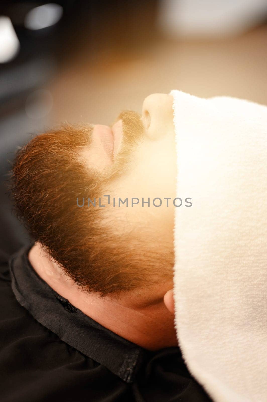 A client is lying on a chair in a barbershop with his beard wrapped in a towel. A towel covers the mans eyes.