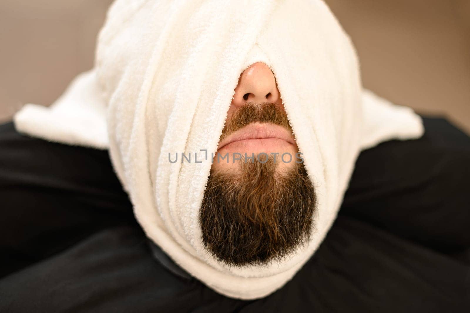 A client is lying on chair in a barbershop with his beard wrapped in a towel. Steaming the beard before the royal shave. by Niko_Cingaryuk