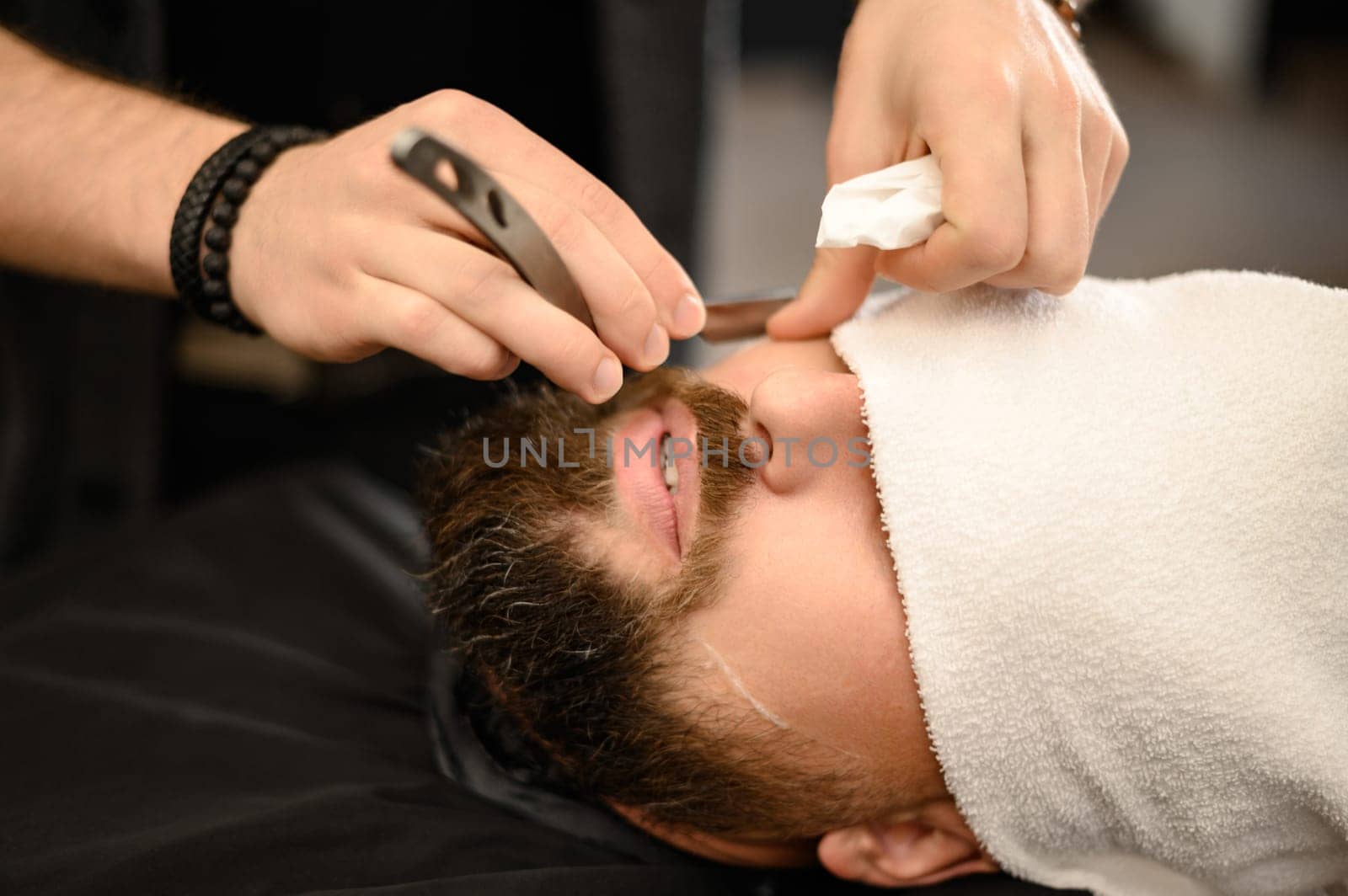 A barber shaves the cheek of a bearded customer with a dangerous razor. Shaving the contour of the beard for the correct shape.