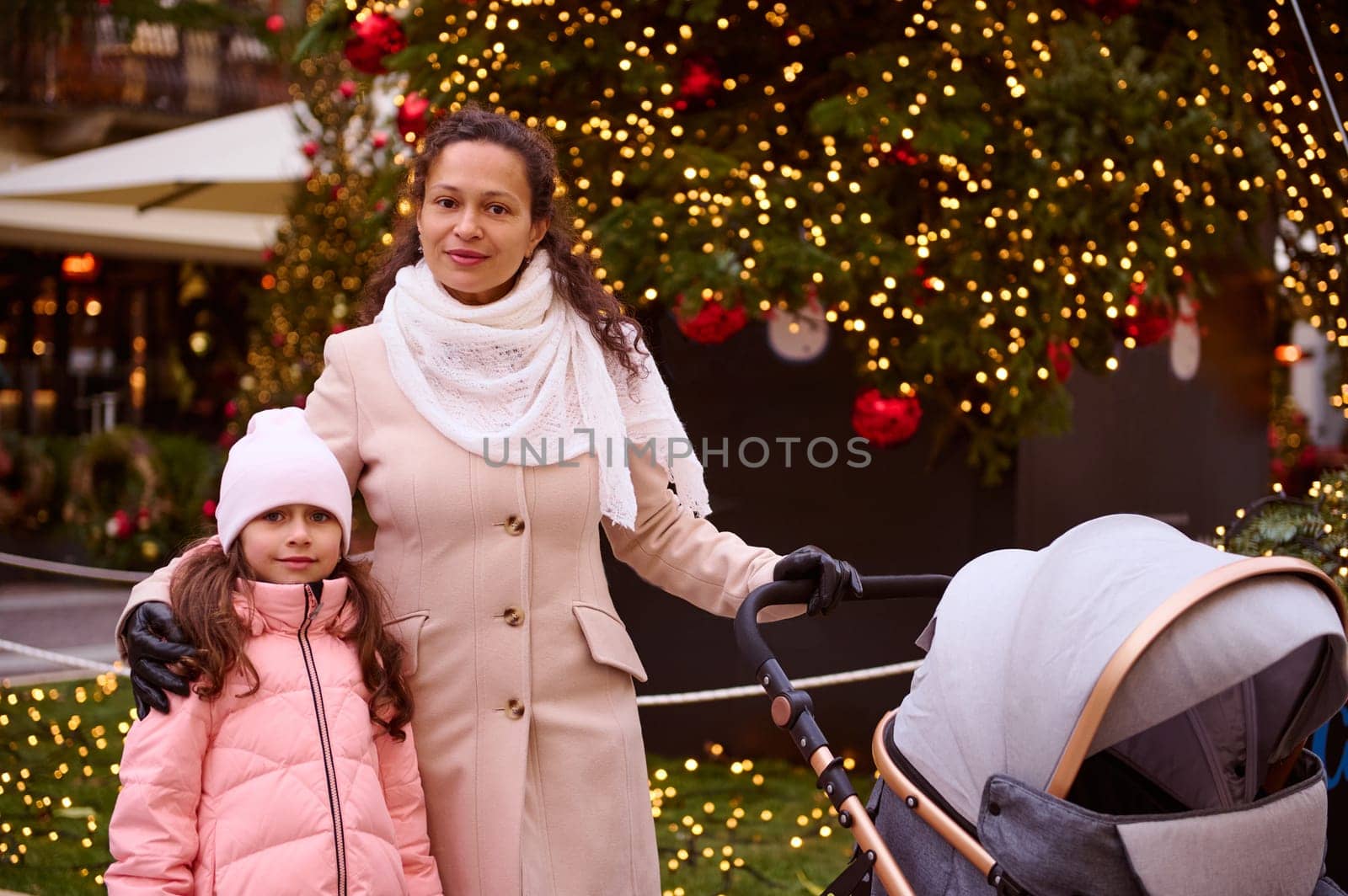 Beautiful family of a multi ethnic happy young mother with her daughter, pushing baby pram, looking at camera, standing against illuminated Christmas tree while strolling the street at Xmas fair