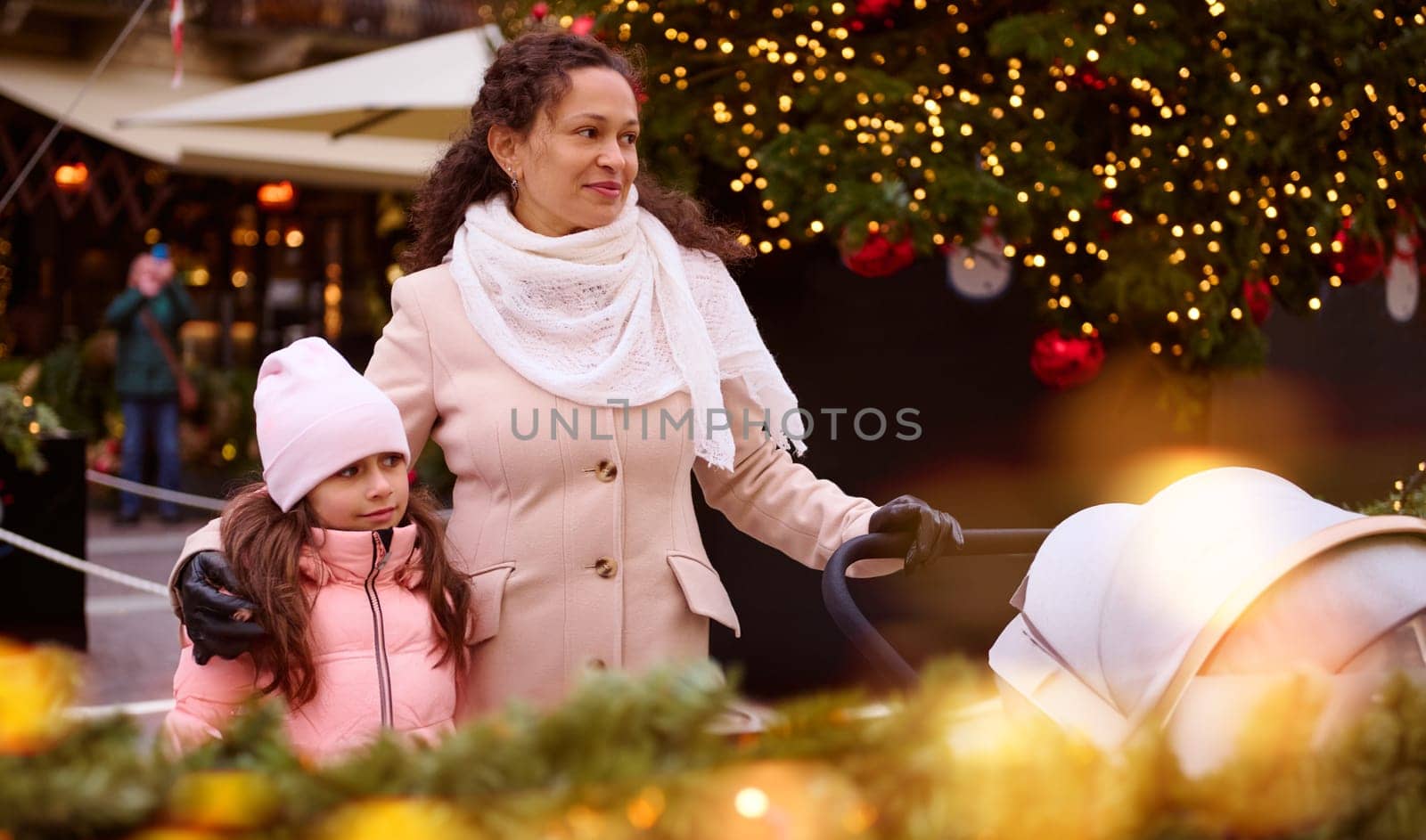 Smiling multi ethnic pretty woman, loving mother hugging her lovely child daughter and pushing baby pram, strolling the Italian street, decorated with garlands and lights with bokeh on the foreground