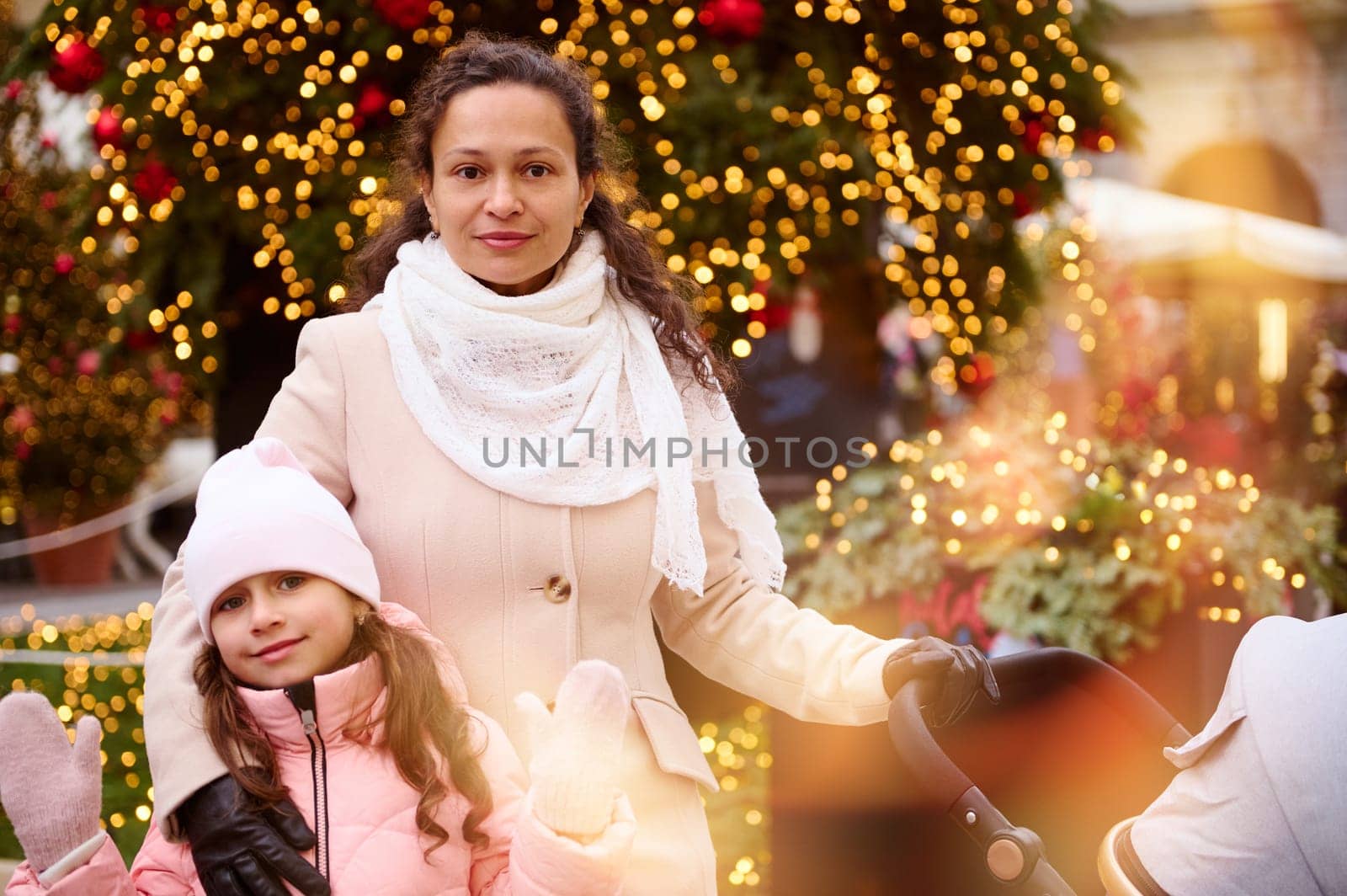 Beautiful mother with baby pram and her cute daughter looking at camera, standing against a Christmas tree outdoors, enjoying festive atmosphere during traditional family market. Bokeh on foreground