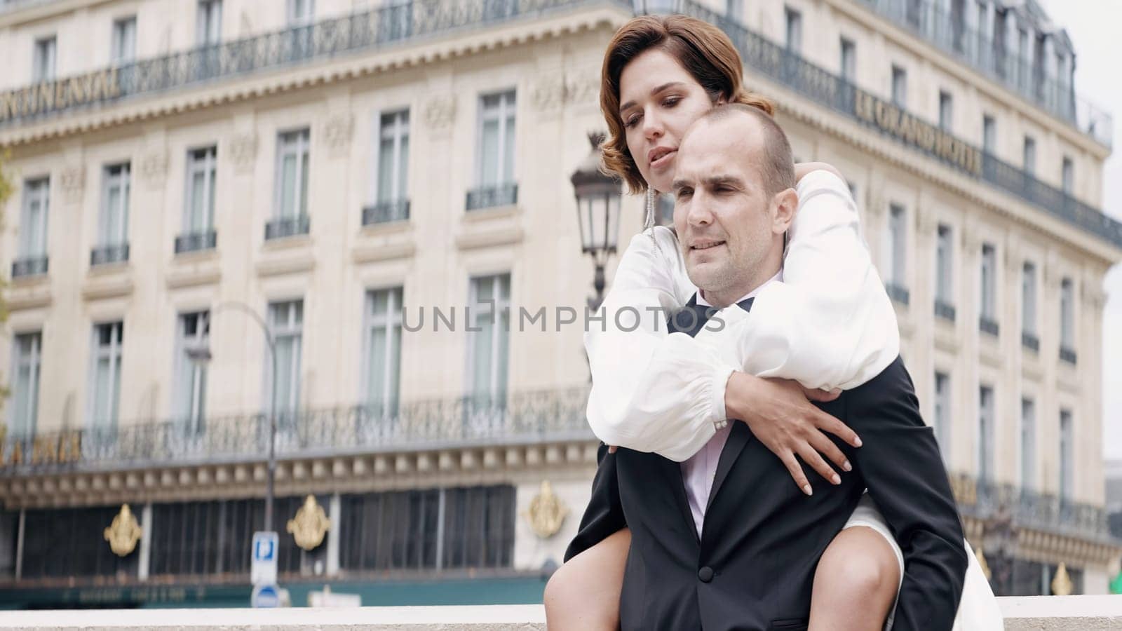 Portrait of a groom carrying happy bride on his back in the city street. Action. Husband and wife newlyweds at the video shooting outdoors