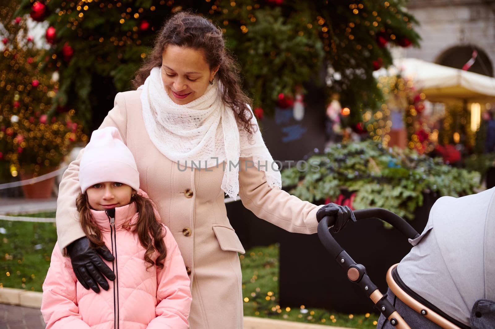 Mother and daughter walking together at Christmas fair. Happy woman hugging her child and pushing baby stroller, enjoying the traditional family market in December. Xmas in Italy. People and holidays