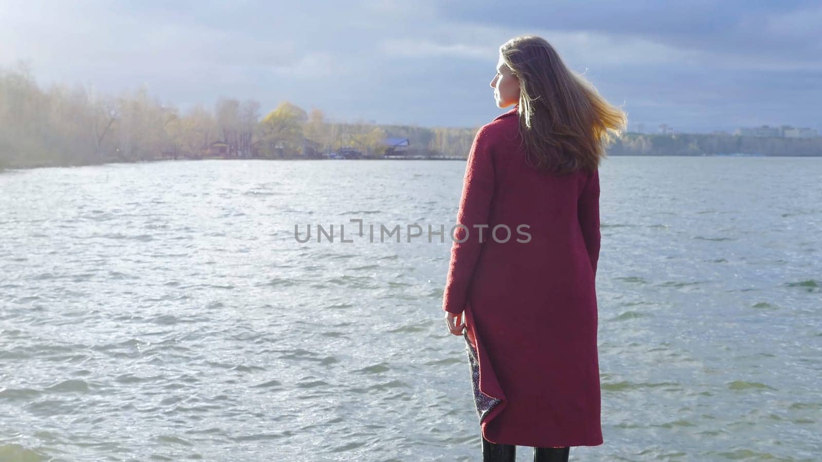 Young girl with her hair stands near the water in the cold season. Back view. Young woman standing near a lake.