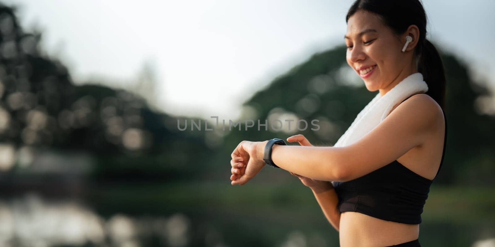 Portrait of smiling Asian woman looking at her smartphone and watch, standing in park wearing sport uniform.