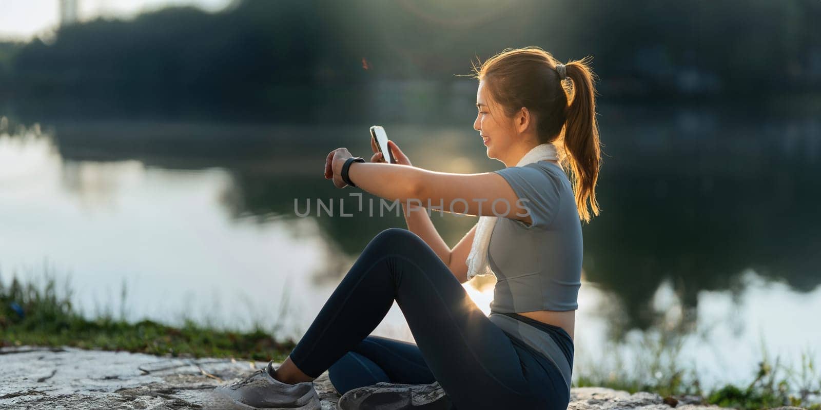 Portrait of smiling Asian woman looking at her smartphone and watch, standing in park wearing sport uniform.