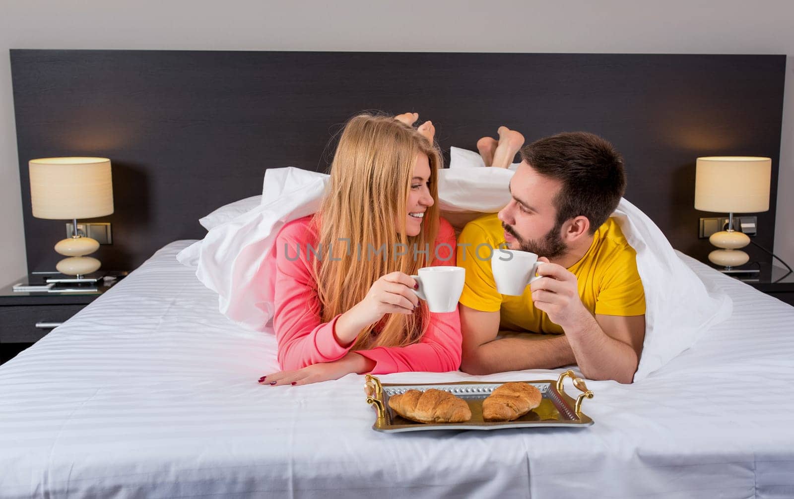 Happy young couple having breakfast tray on bed at home in bedroom. Couple drink coffee in bed, happy smile