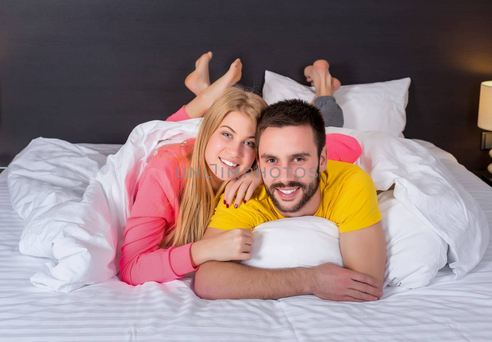 Young lovely couple lying and have fun in a bed, happy smile looking at camera, cover under blanket.
