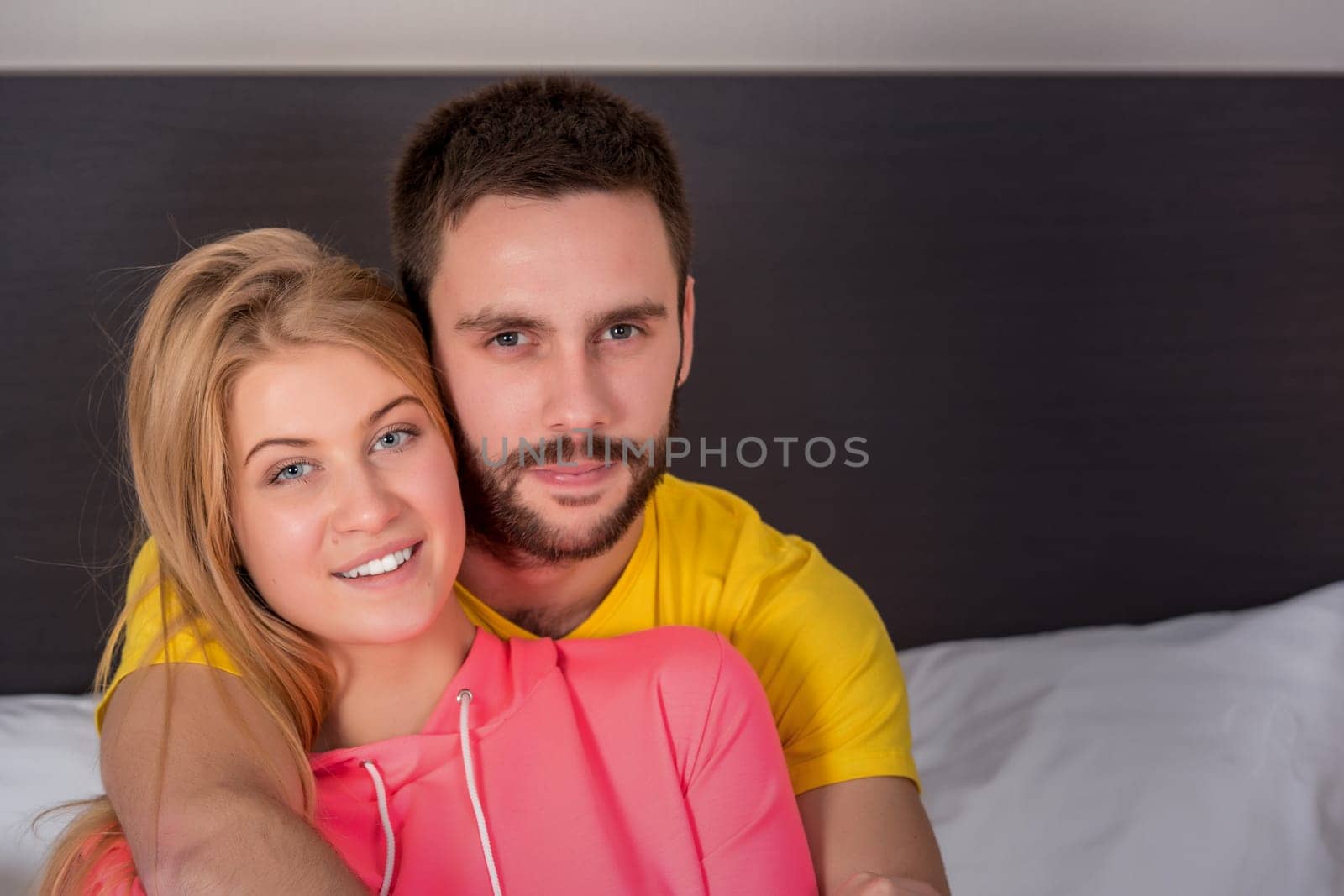 Young lovely couple have fun in a bed. Woman hugging man, happy smiles looking at camera