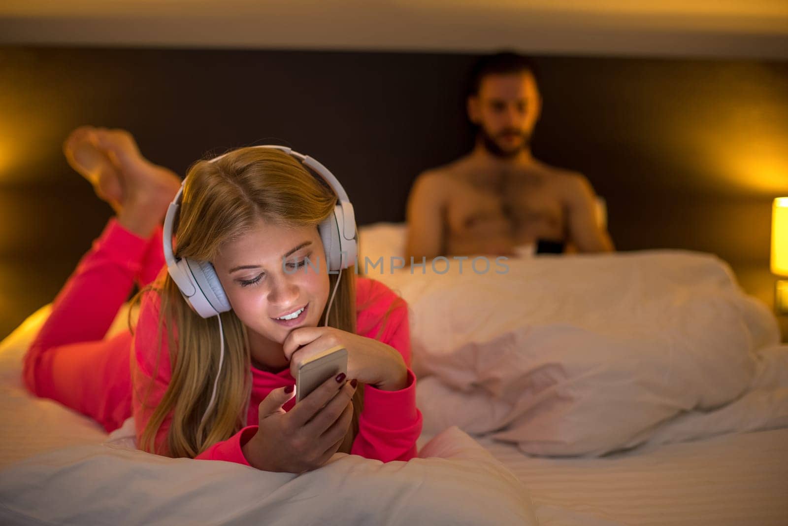 Young happy women on white bed using mobile phone with headphones, boyfriend on background. Concept about technology and people