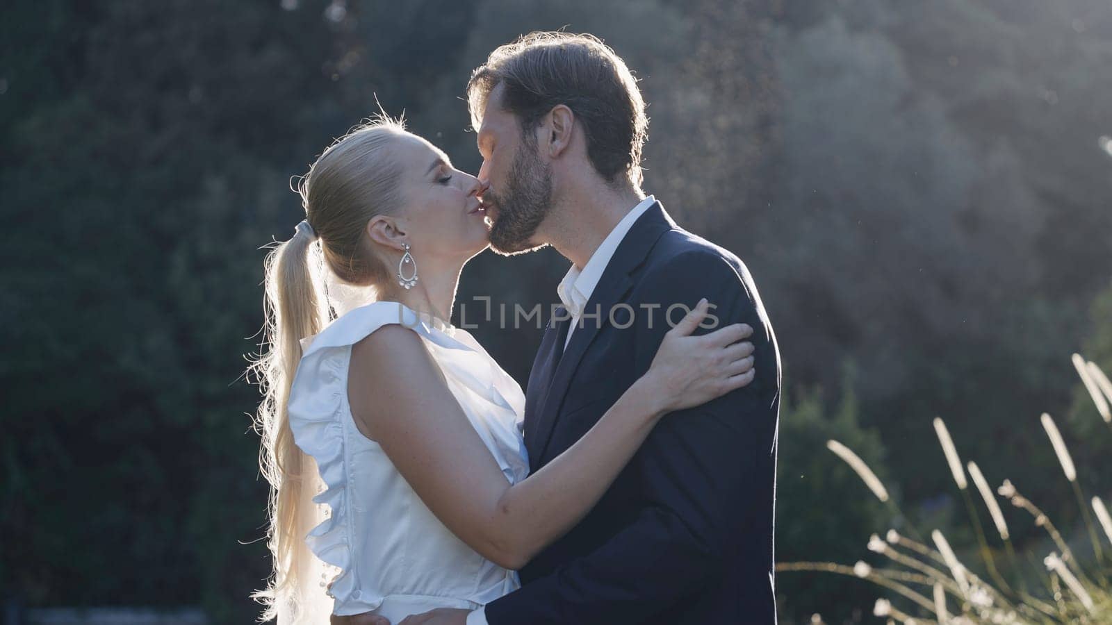 Newlyweds kiss in nature with sunlight. Action. Beautiful couple of newlyweds in suits are kissing. Elegant couple of lovers kissing on background of sunlight and nature.