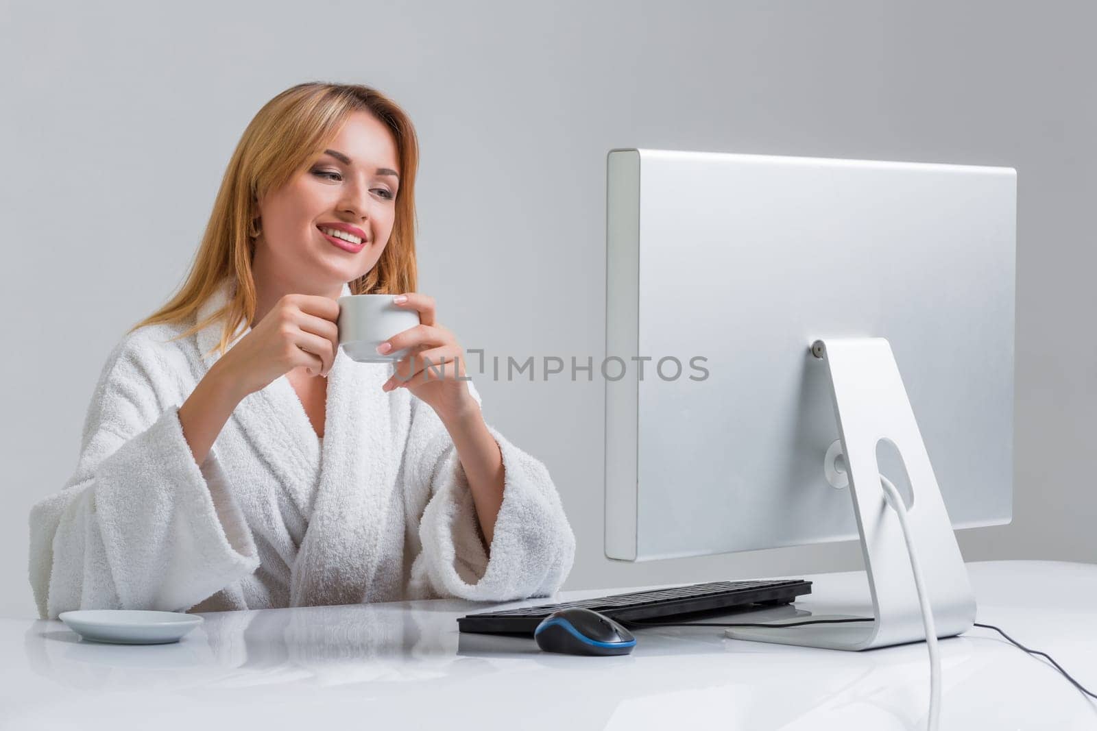 young woman sitting in the table and using computer on gray background. girl smiling and looking at monitor. with a cup in hands