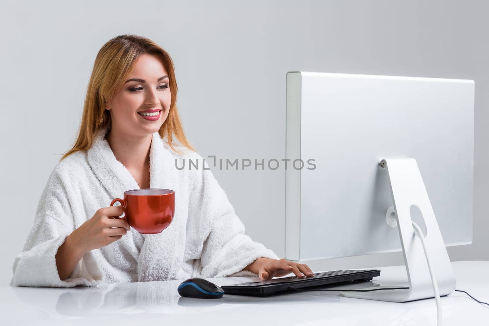 young woman sitting in the table and using computer on gray background. girl smiling and looking at monitor. with a cup in hands