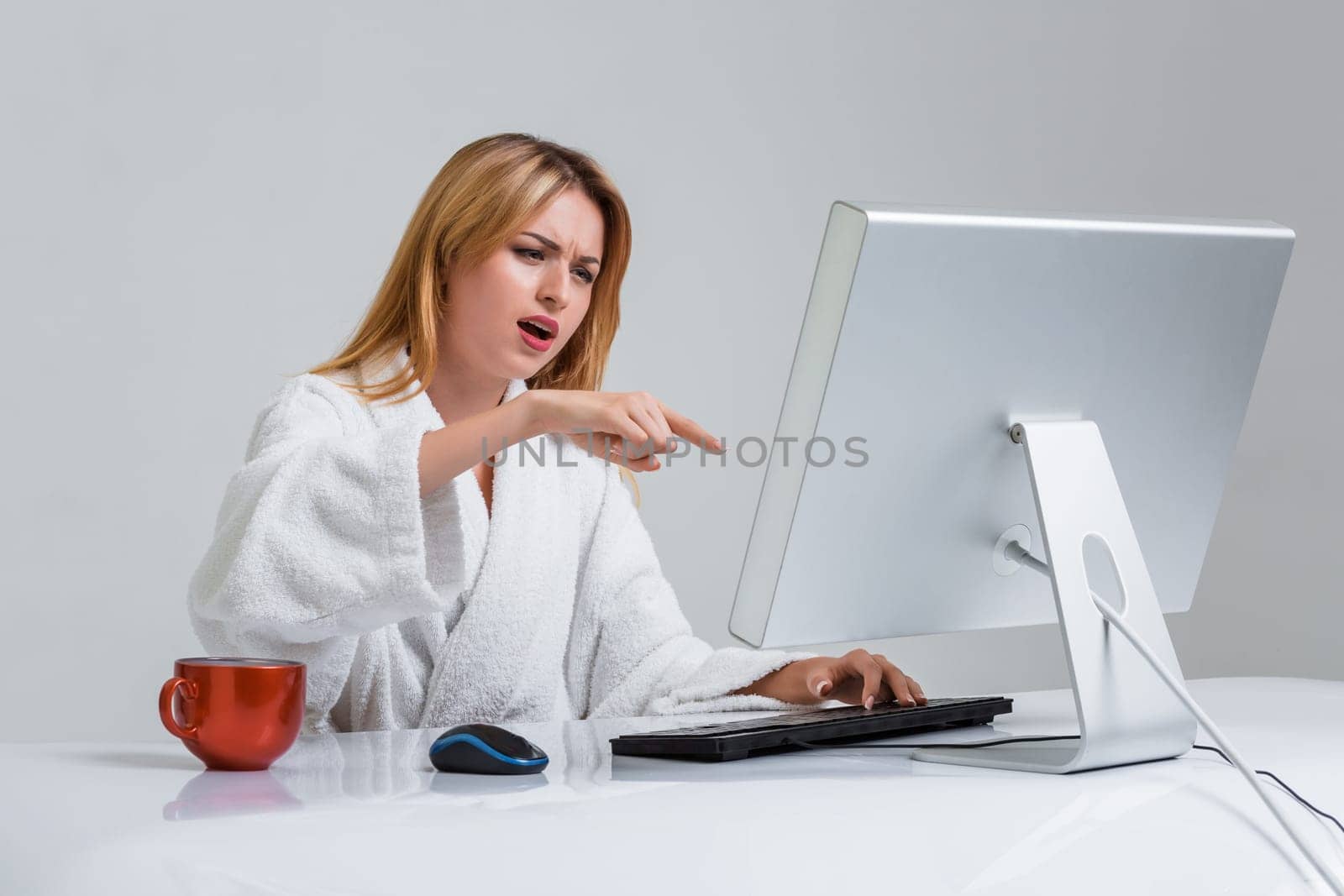 young woman sitting in the table and using computer on gray background. indignation is pointing at the monitor