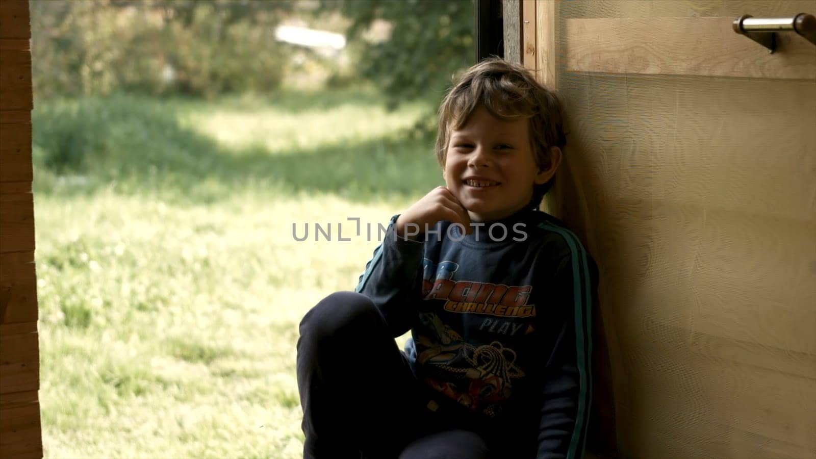 Cute boy laughing at door. Creative. Happy boy is sitting on porch of country house. Boy is resting and laughing on background of cottage yard.