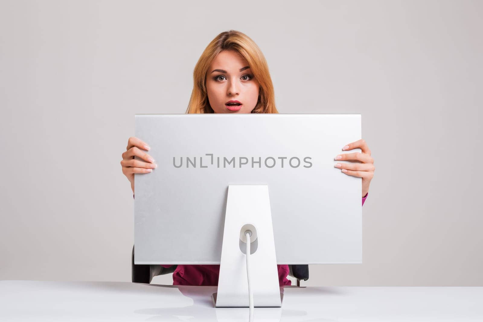 young woman sitting in the table and using computer on gray background. Surprised and scared hidden behind the monitor. It peeping from behind him