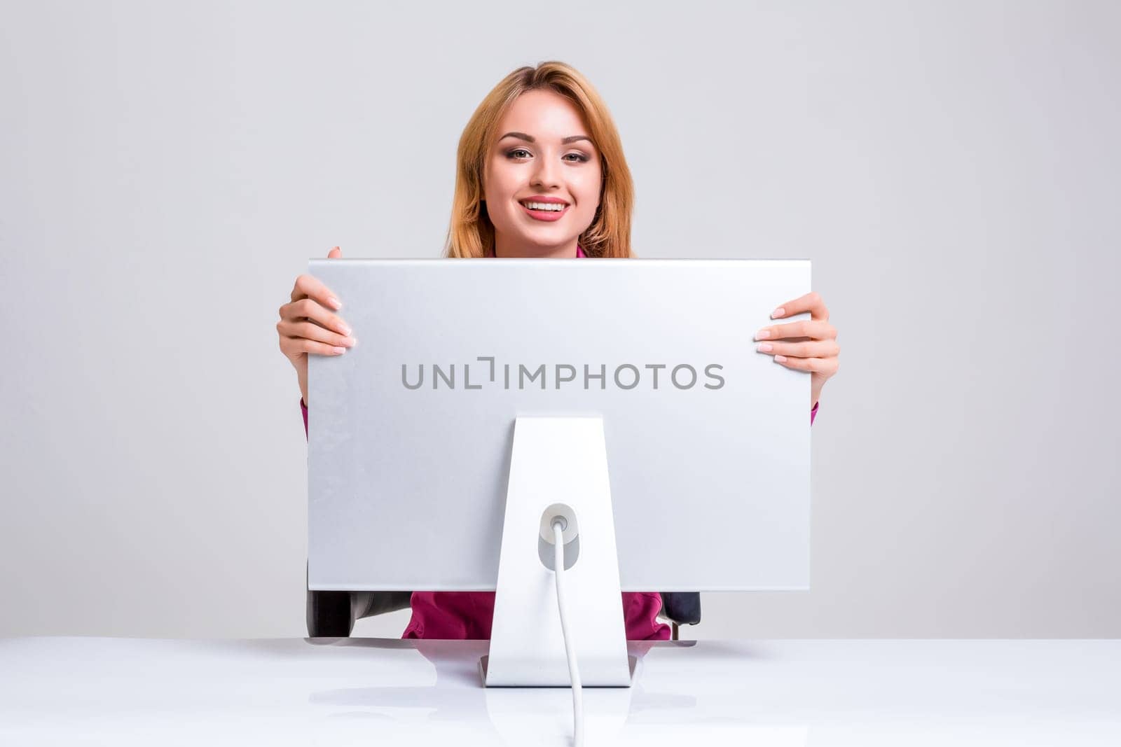 young woman sitting in the table and using computer on gray background. Surprised and scared hidden behind the monitor. It peeping from behind him