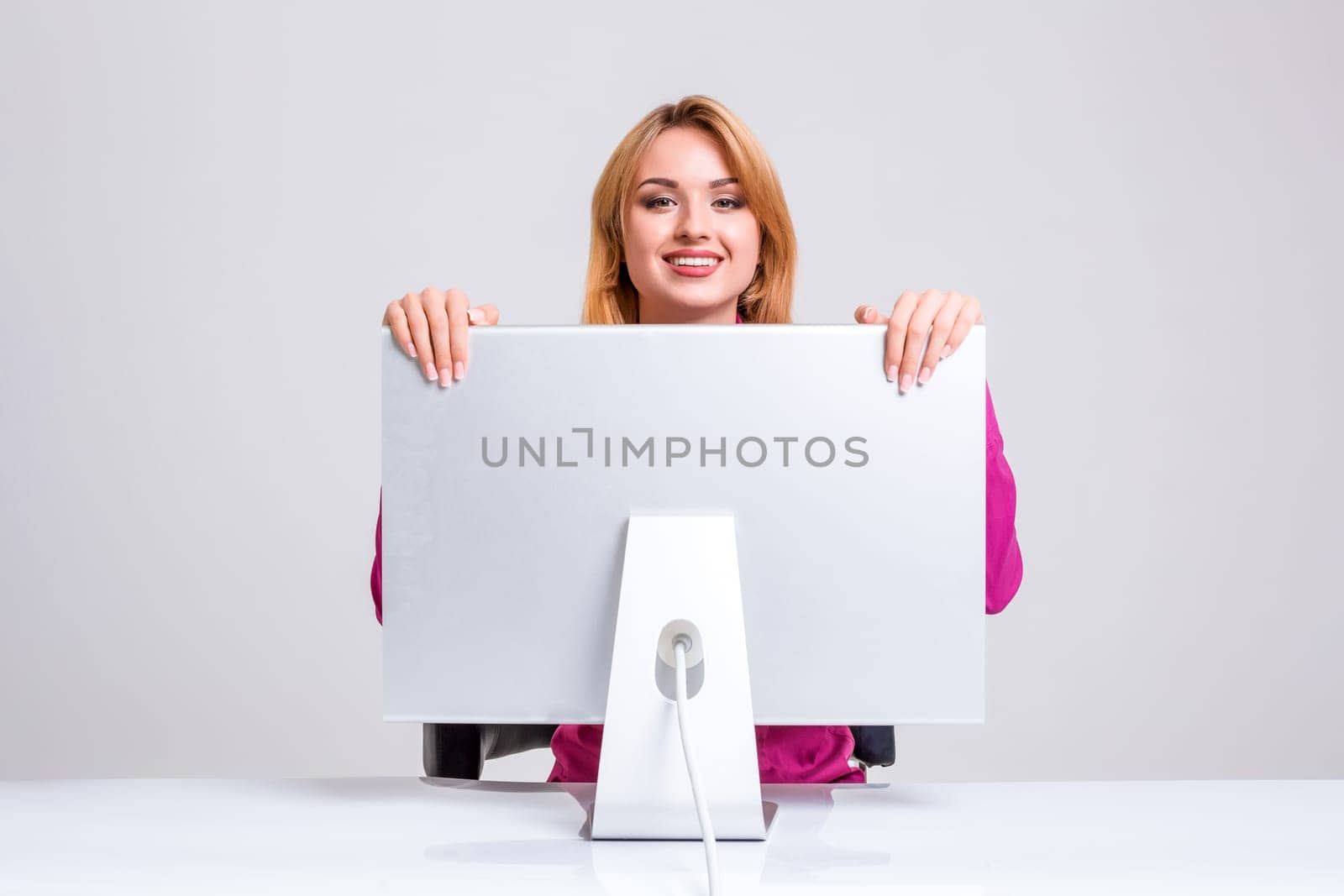 young woman sitting in the table and using computer on gray background. Surprised and scared hidden behind the monitor. It peeping from behind him