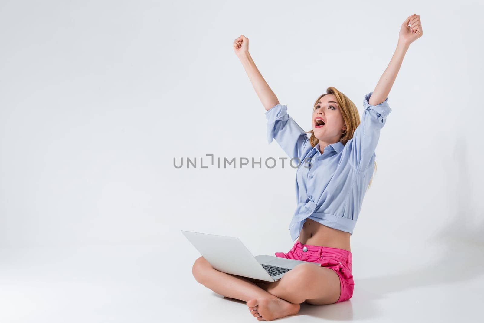 young woman sitting on the floor with crossed legs and using laptop on white background. very satisfied, happy, she raised her hands up