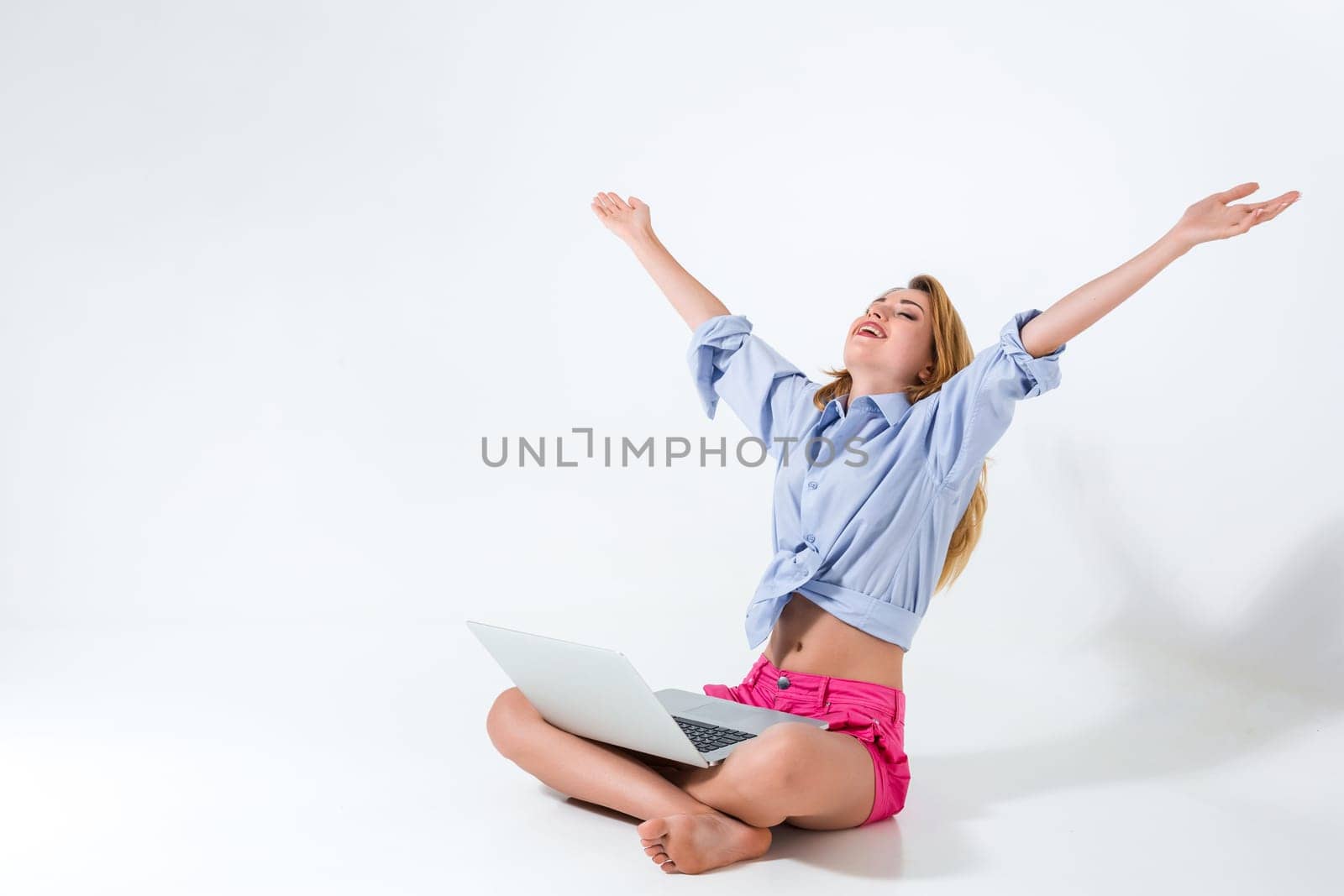 young woman sitting on the floor with crossed legs and using laptop on white background. very satisfied, happy, she raised her hands up